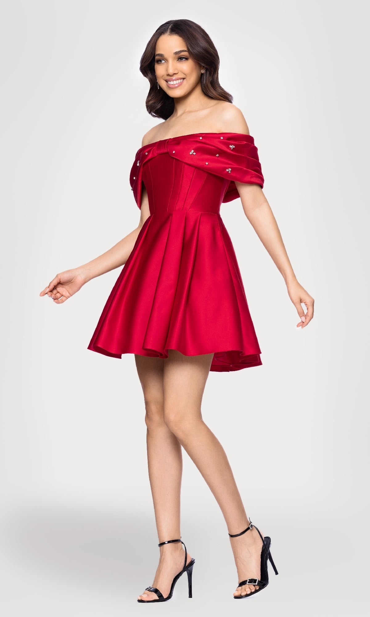 A woman models the Off-Shoulder Red Hoco Dress: Blondie Nites 5695BN, featuring a flared skirt, paired with black high-heeled sandals, standing against a plain light background.